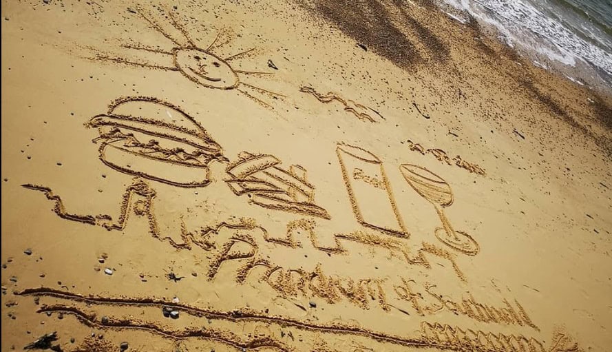 Sand art of a burger, drink, sun, and ocean waves on the beach.