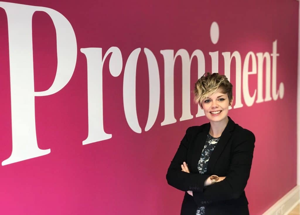 Professional woman standing in front of "Prominent" sign at Suffolk Business event.