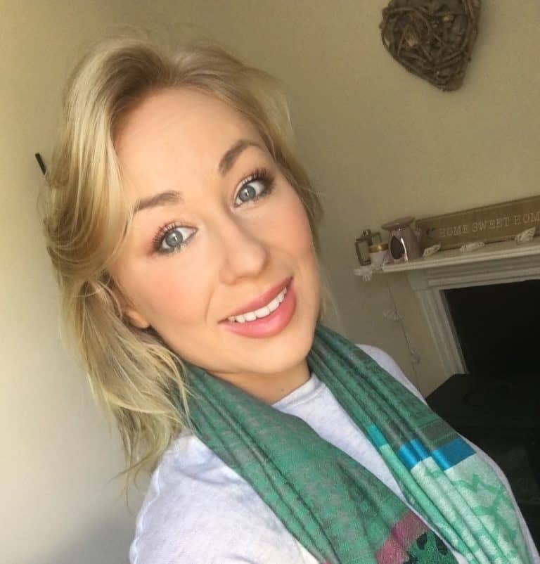 Portrait of a smiling young woman with blonde hair and blue eyes, wearing a green scarf indoors.