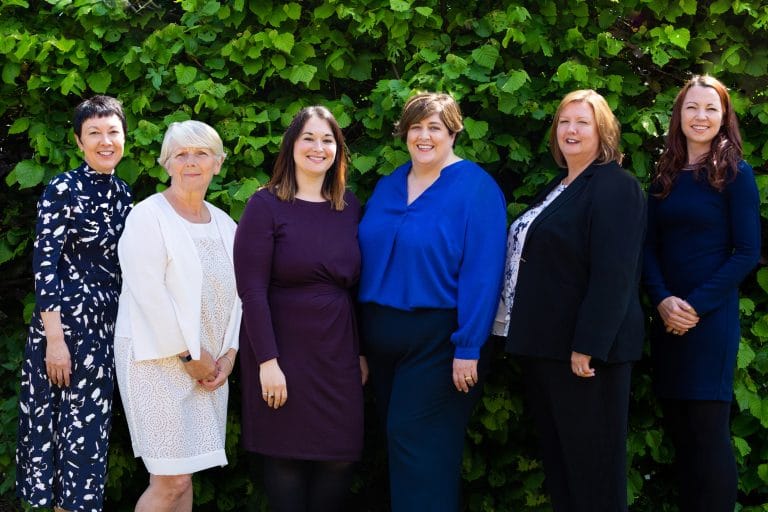 Suffolk Business women team standing outdoors in front of green foliage.
