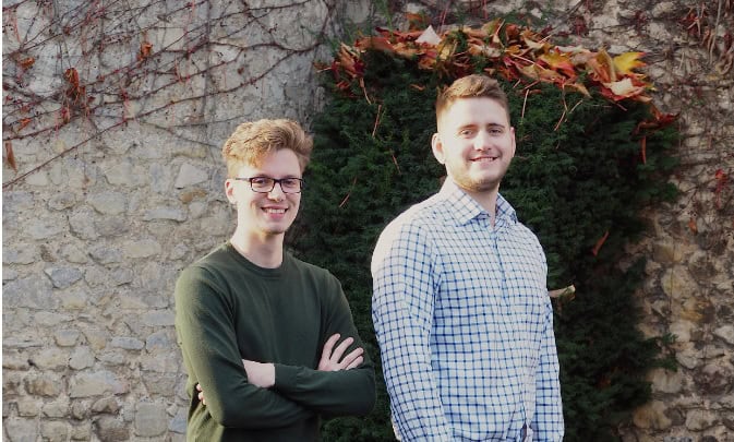 Two young men smiling outdoors in front of a stone wall and autumn foliage.