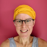 Smiling woman with glasses and yellow headscarf against pink background.