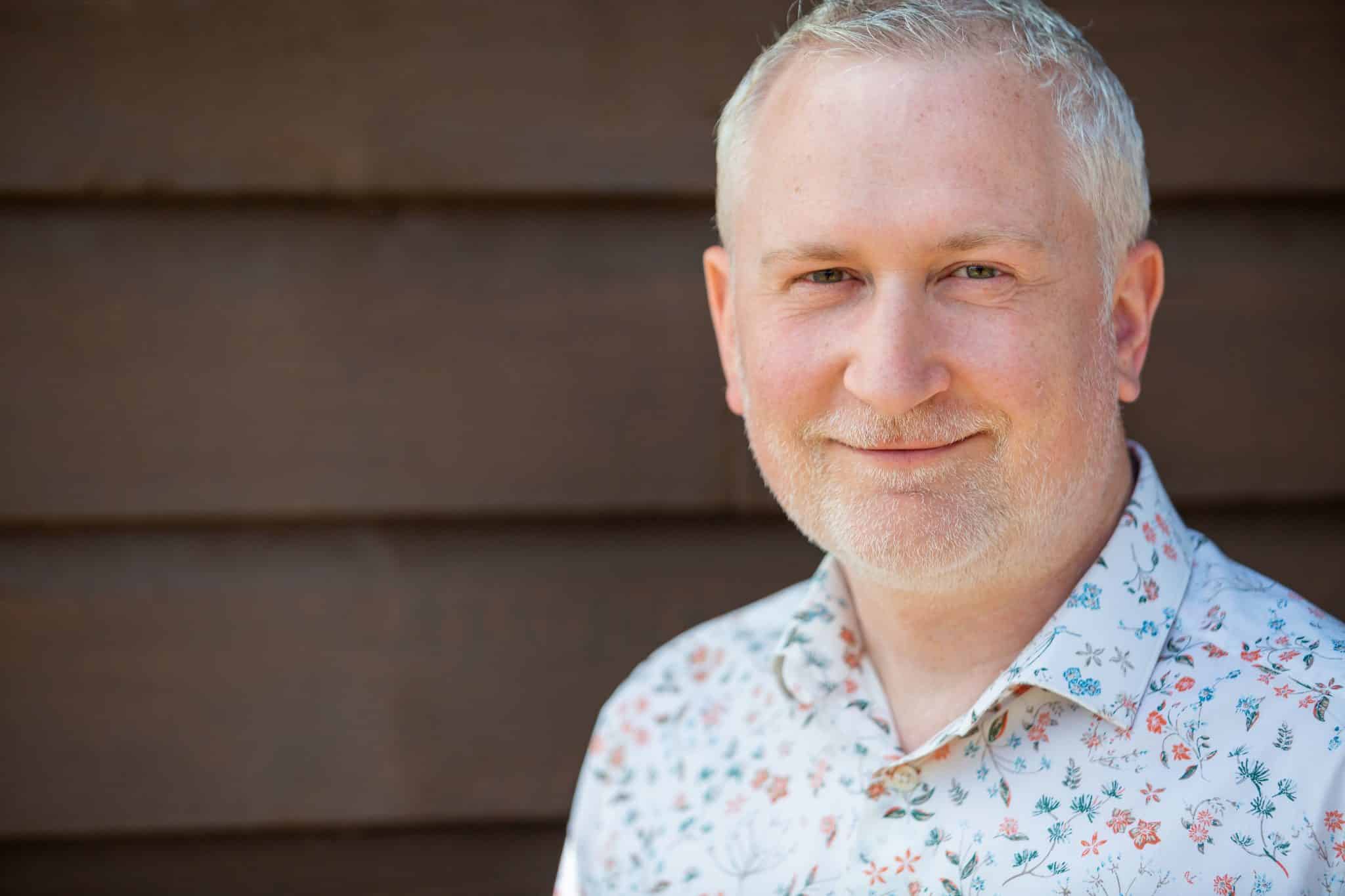 Portrait of a smiling man in a floral shirt against a wooden background.