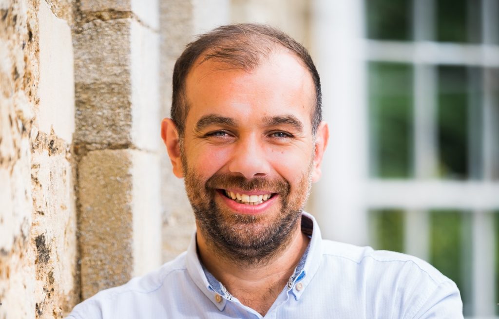 Smiling businessman in Suffolk office with brick wall and window background.