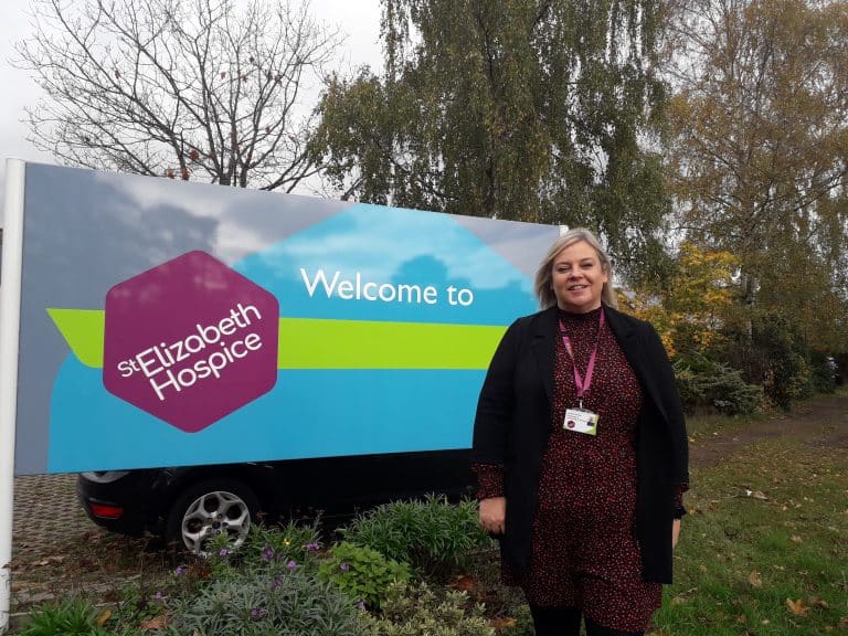 Volunteer at Suffolk Hospice standing outdoors near welcome sign.