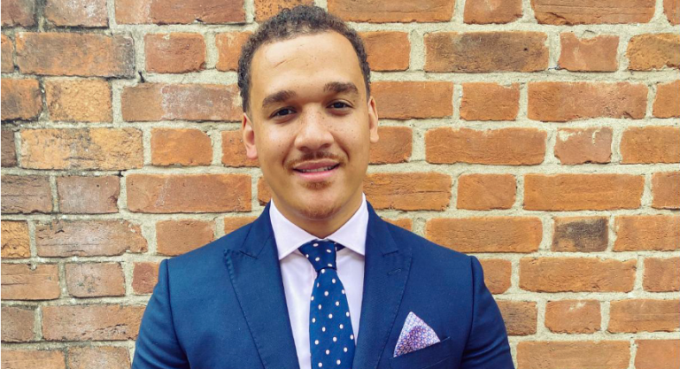 A young professional man in a blue suit and polka dot tie standing against a brick wall.
