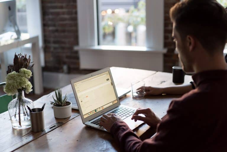 Man working on a laptop at a home office desk with plants and a coffee mug.