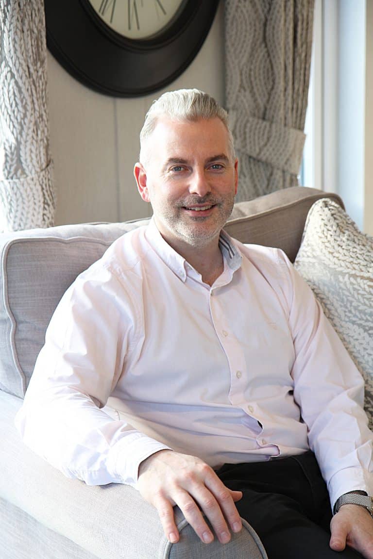Mature man in white shirt sitting on sofa in Suffolk home, smiling at camera.