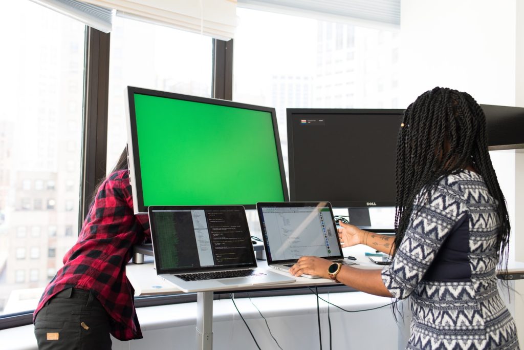 Two professionals collaborating with multiple monitors and laptops in a modern office setting.