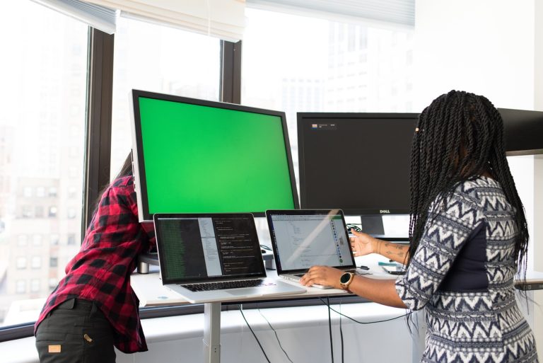 Two professionals collaborating with multiple monitors and laptops in a modern office setting.