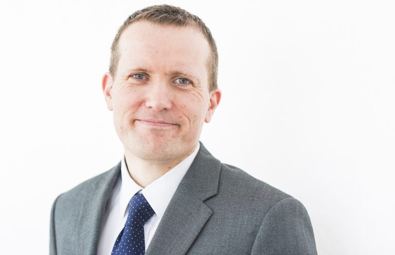 Business professional portrait of a man in a grey suit and blue tie, smiling confidently against a w.
