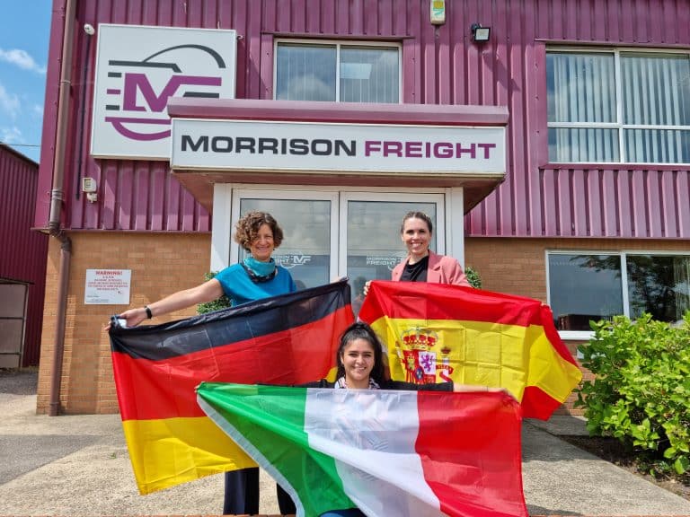 Morrison Freight UK team holding German, Spanish, and Italian flags outside warehouse.