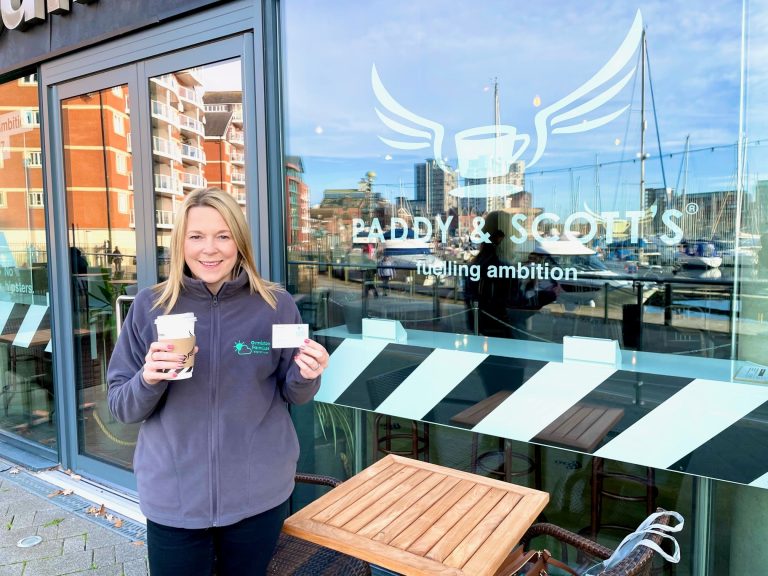 Smiling woman holding coffee and business card outside Suffolk Business store in Ipswich.