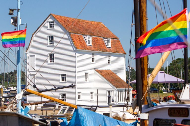 A colourful scene of Suffolk harbour featuring rainbow pride flags flying near boats and a historic.