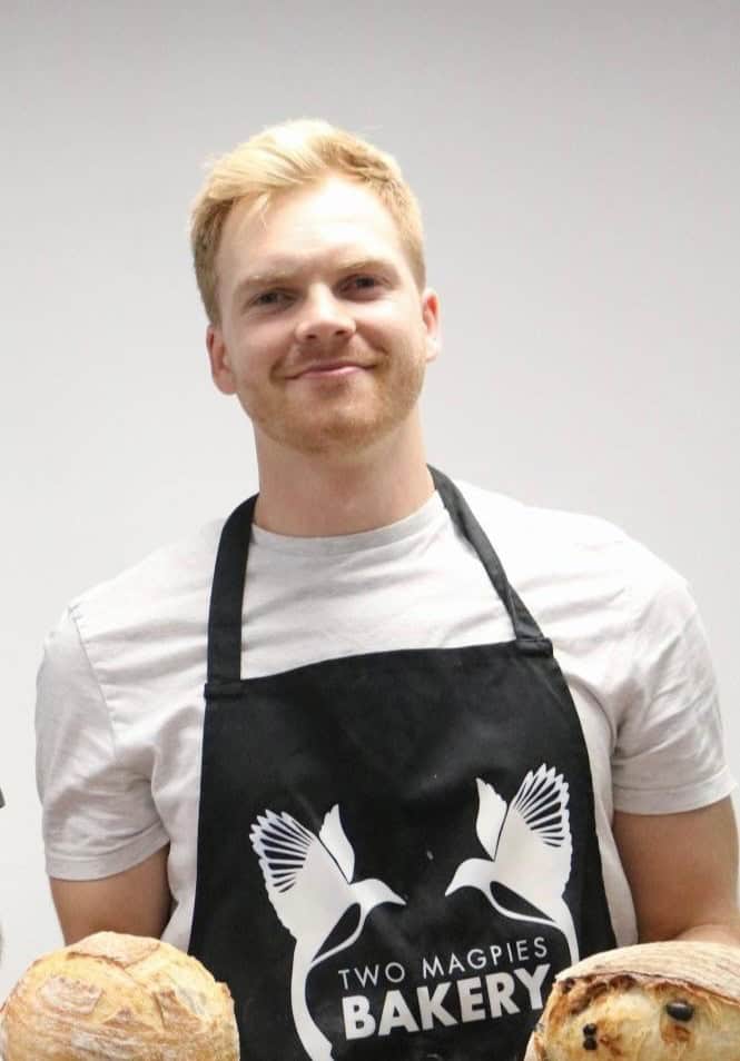 Friendly young man wearing bakery apron with bread, smiling at camera.